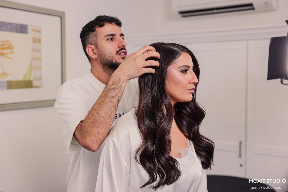 A hairstylist carefully styling a woman's dark wavy hair in a bright salon setting.