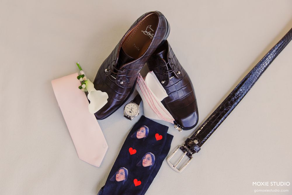 Dark brown oxford shoes with heart-patterned socks, pink tie, belt, and boutonniere arranged as groom wedding accessories.