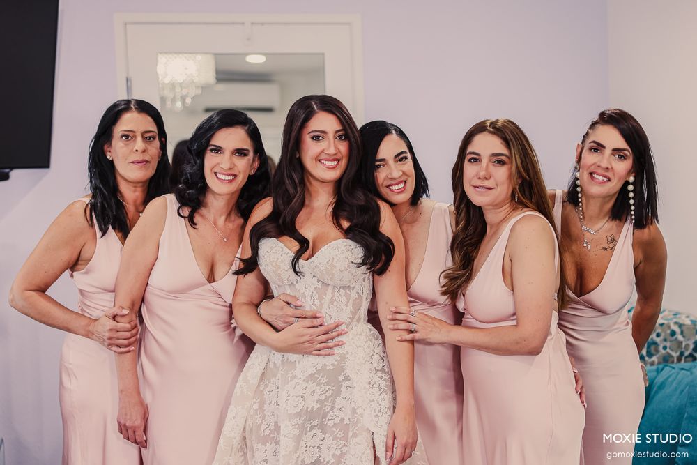 Bride in white lace gown poses with five bridesmaids in blush pink dresses during wedding day preparations.