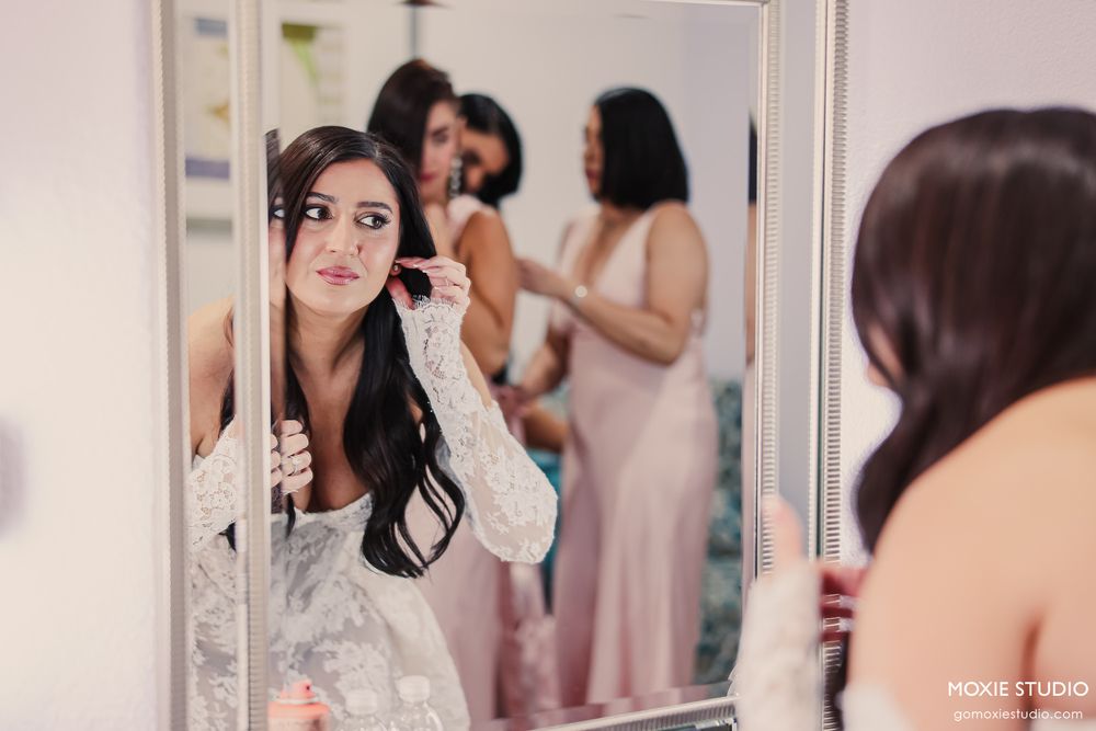 Bride checking her earring in a mirror while bridesmaids in pink dresses help her get ready.