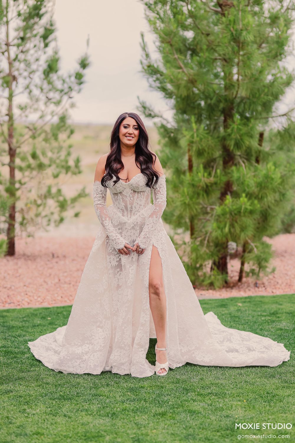 Bride wearing long-sleeve lace wedding dress with high slit posing outdoors among pine trees.