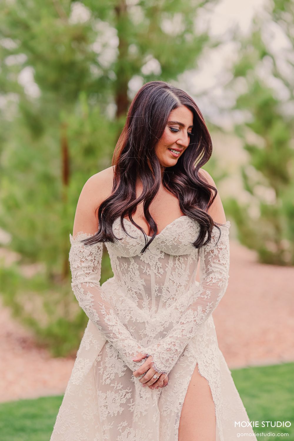 Bride in off-shoulder lace wedding dress looking down while standing outdoors near pine trees.
