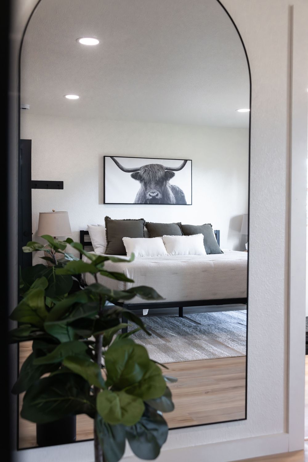 Arched floor mirror reflecting a cozy bedroom with a highland cow art print above a neutral-toned bed.