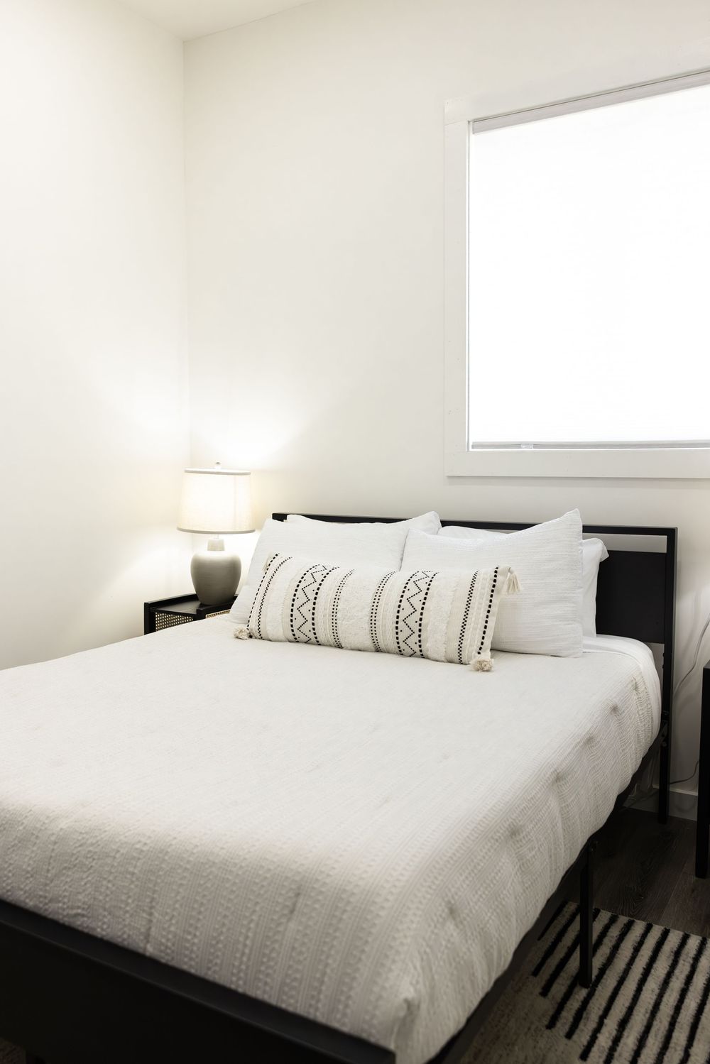 Minimalist bedroom with black bed frame, white bedding, decorative pillows, nightstand lamp, and bright window.