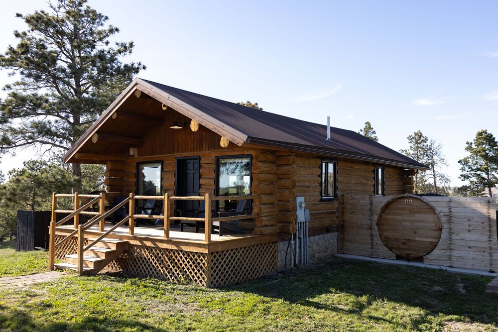 Rustic log cabin with covered deck, brown metal roof, and large barrel feature surrounded by pine trees.