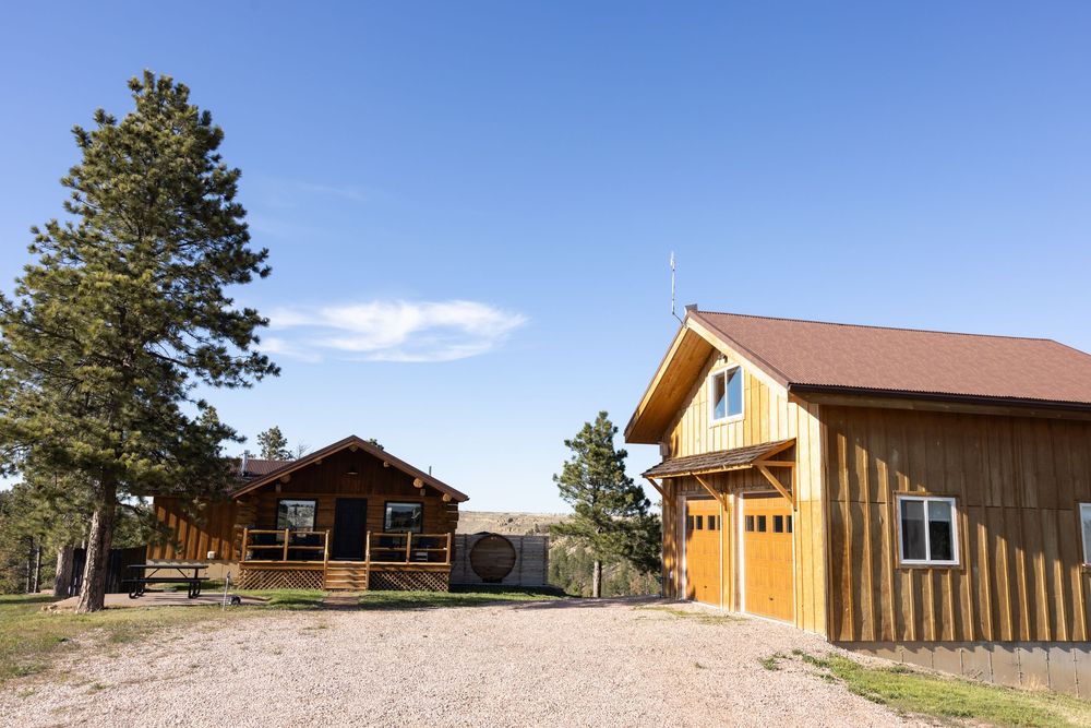Two rustic wooden cabins with gravel driveway surrounded by pine trees under a clear blue sky in a rural mountain setting.