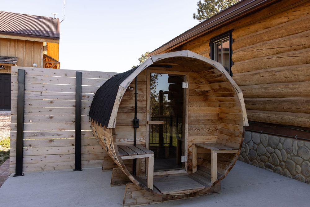 Rustic barrel sauna with wooden exterior and glass door beside a log cabin on a concrete patio.