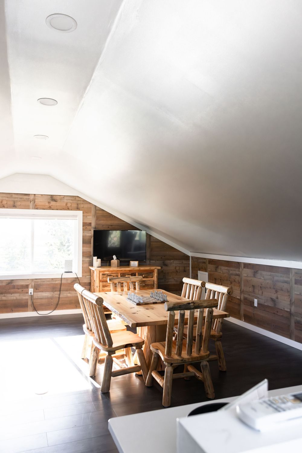 Rustic attic dining room with wooden walls, log chairs, farmhouse table, dark hardwood floors, and wall-mounted TV.