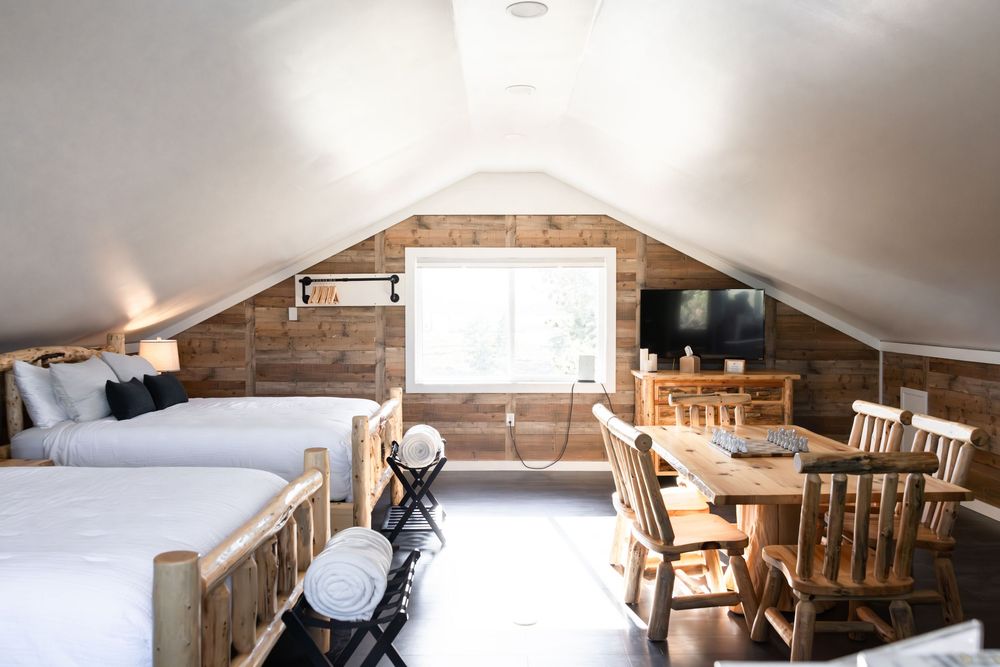 Rustic attic bedroom with vaulted ceilings, wood-paneled walls, two log-frame beds, and a wooden dining set near a window.
