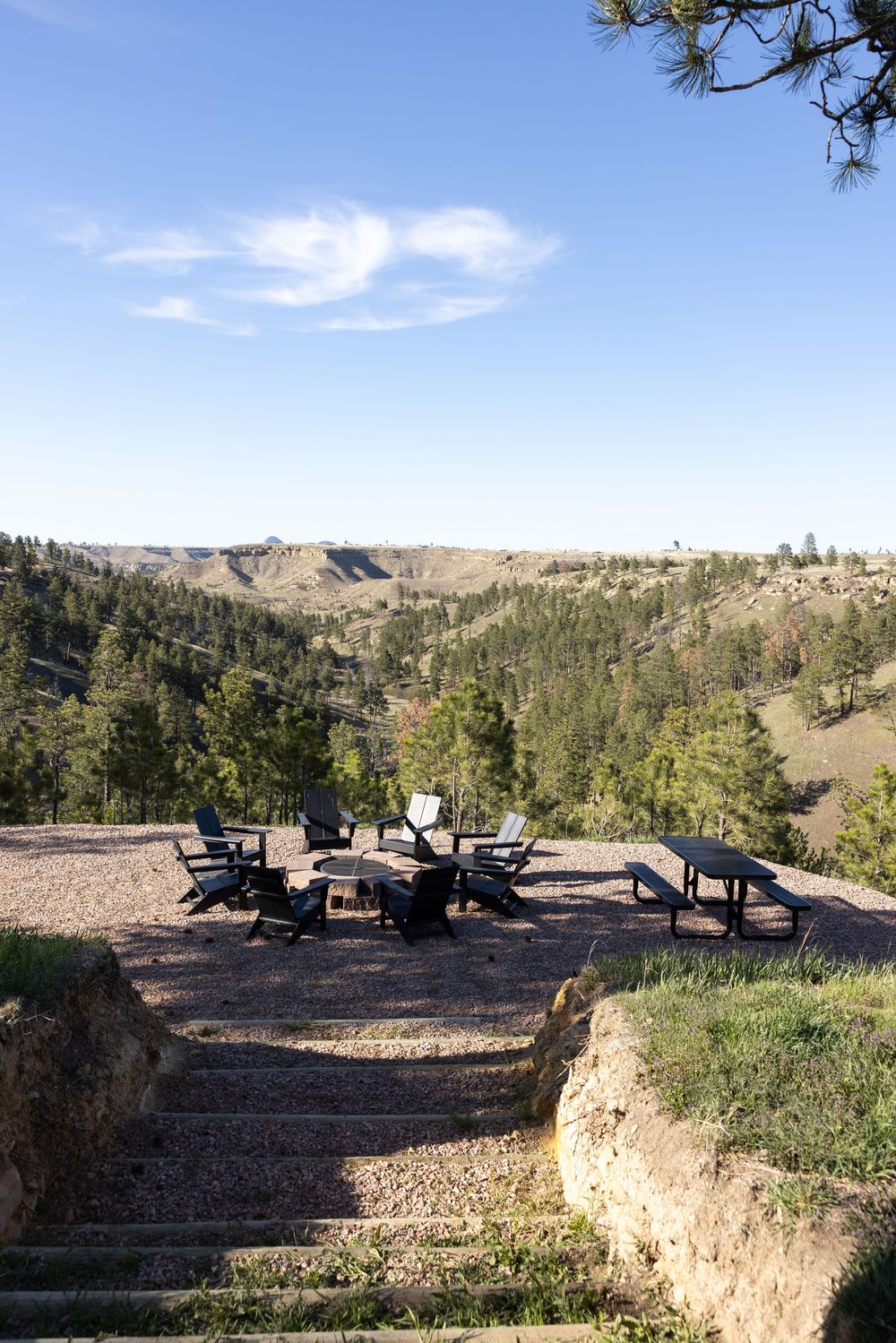 Outdoor fire pit with Adirondack chairs on a gravel patio overlooking a forested canyon under a clear blue sky.