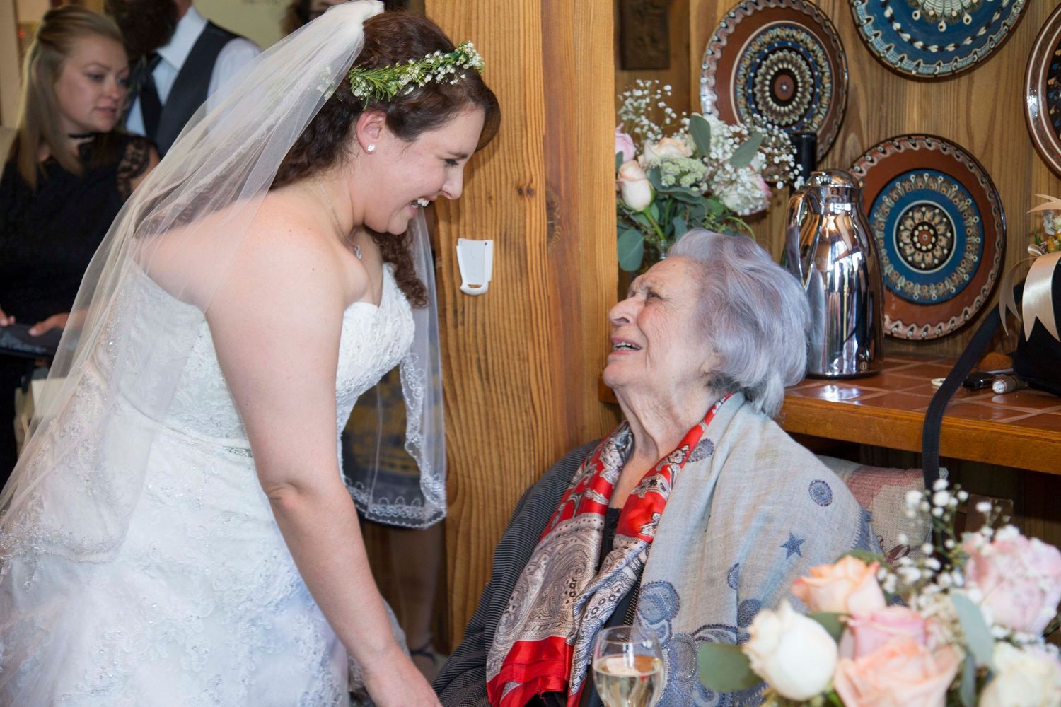 Bride in white gown smiles warmly at elderly seated woman holding champagne at a wedding reception.