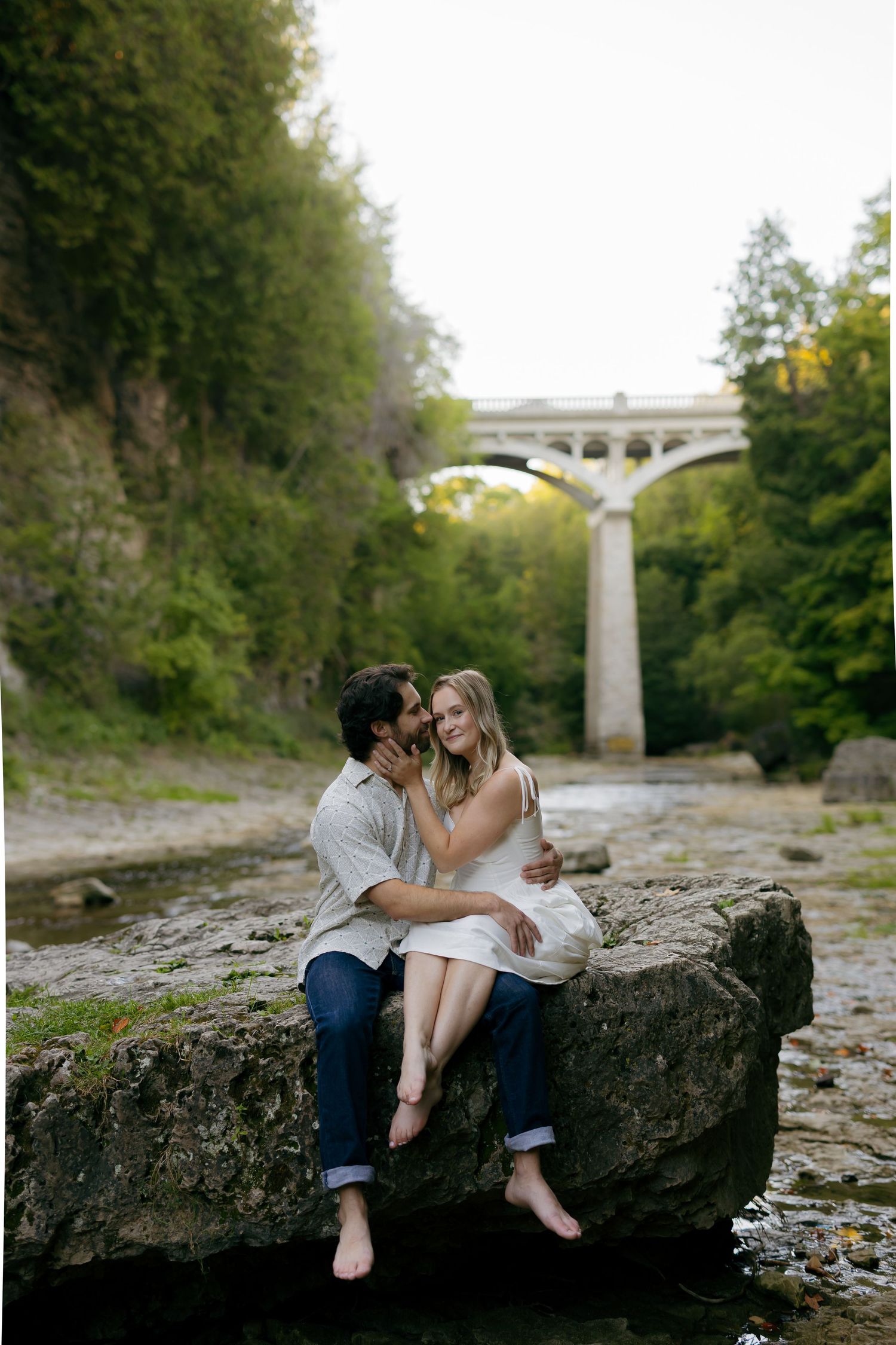 Couple sitting barefoot on a rock by a creek, embracing romantically with a stone arch bridge and lush greenery behind them.
