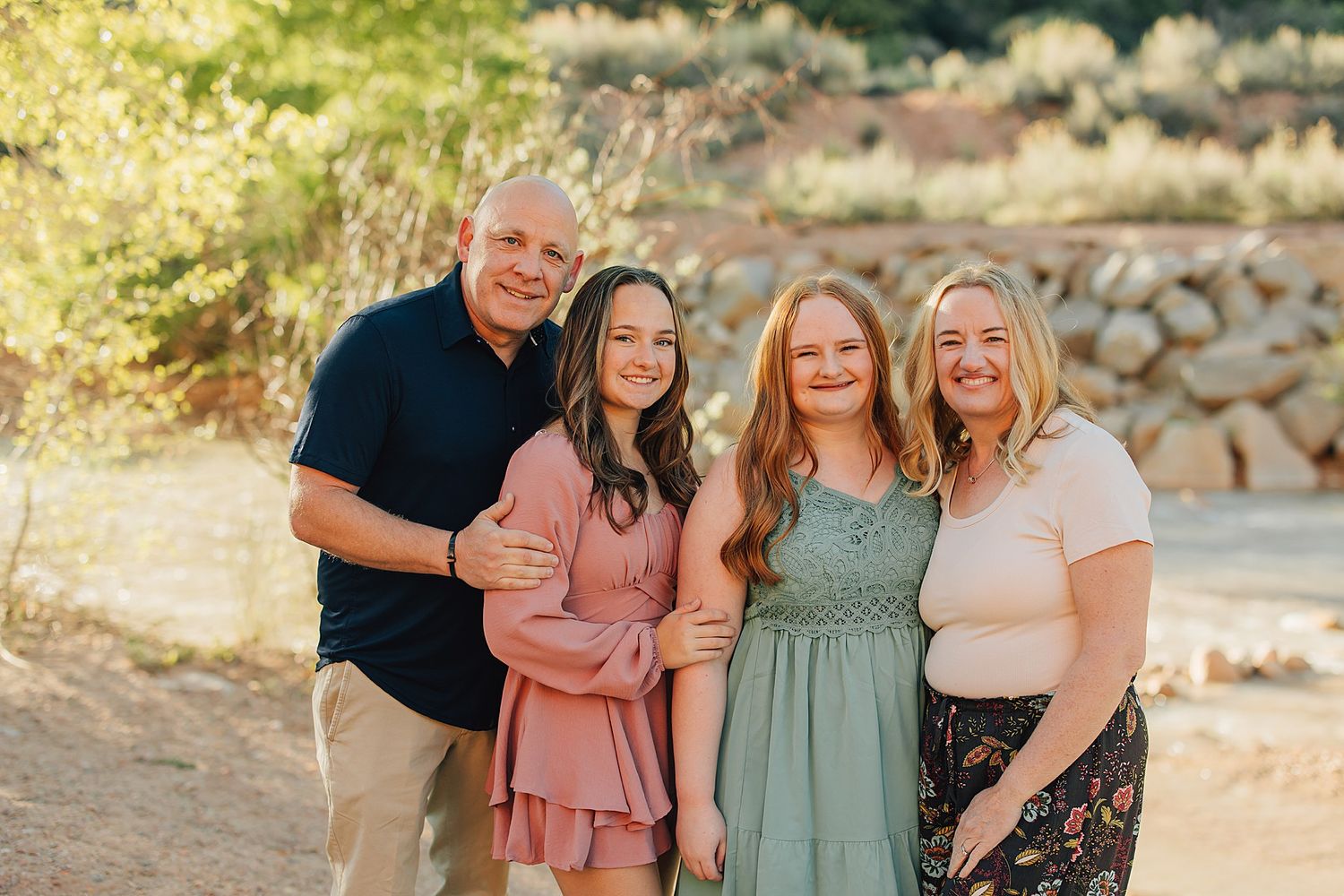 Family of four smiles together outdoors, two teenage daughters in pink and green dresses with parents in natural setting.