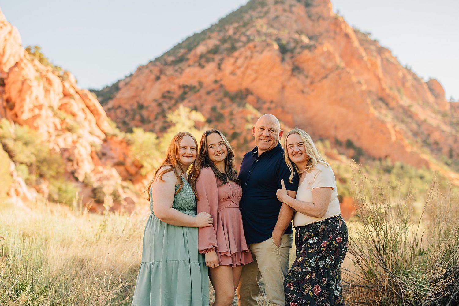 Family of four smiling together in front of red rock mountains in a scenic outdoor desert landscape.