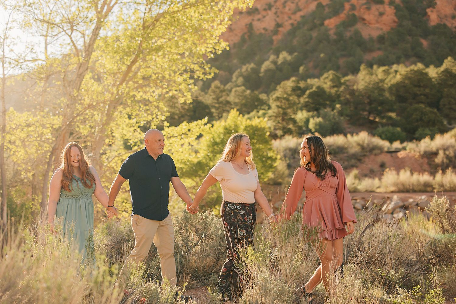 Family of four holding hands walking through a sunlit desert landscape with red rock mountains in the background.