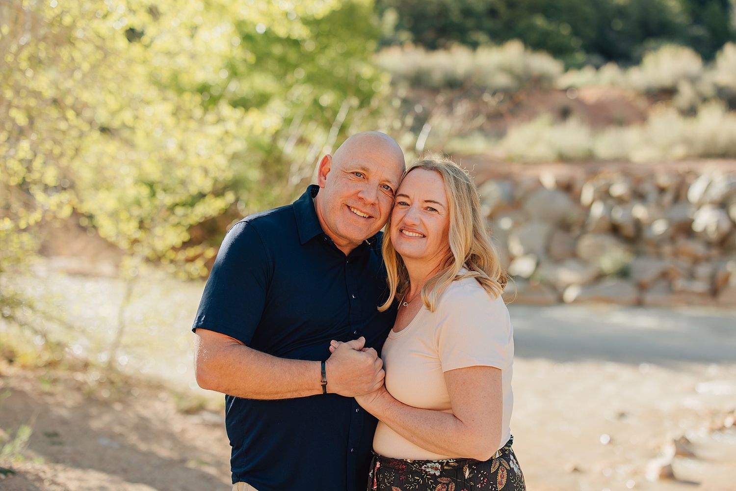 A smiling couple poses outdoors in a sunny natural setting with trees and rocks in the background.