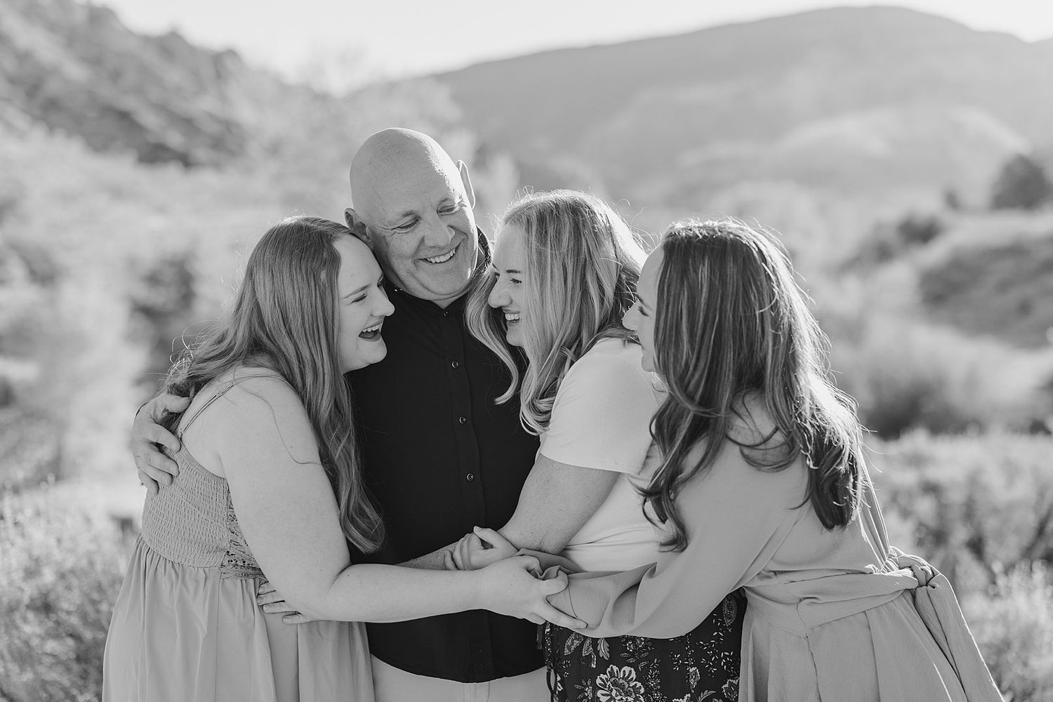 Black and white family portrait of a bald man embracing three daughters outdoors in a mountain setting.