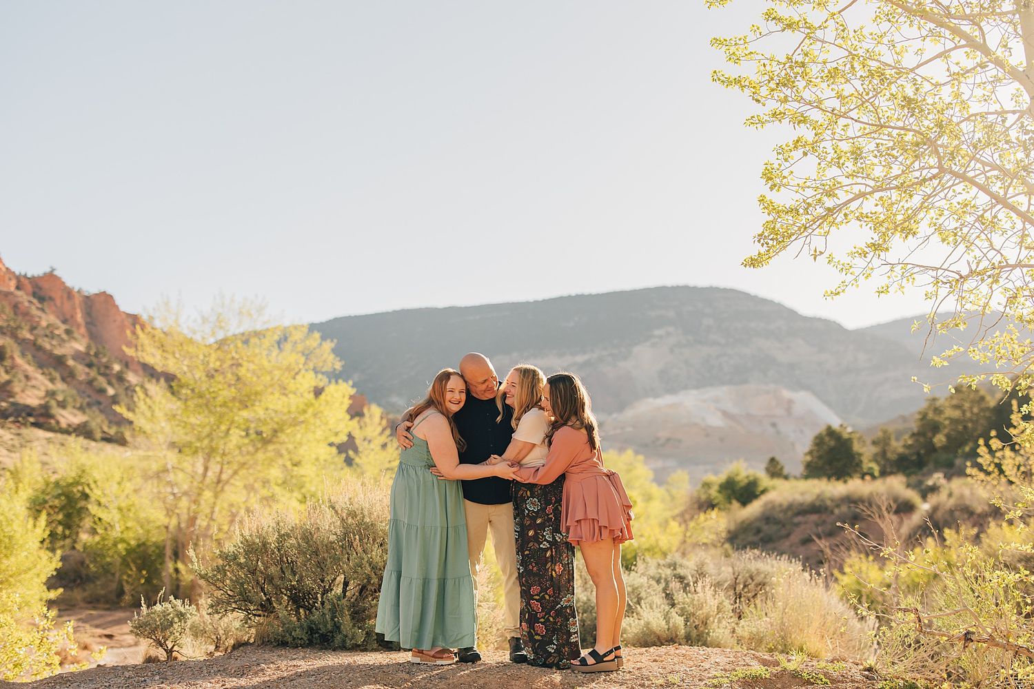 Four women embracing outdoors near red rock mountains with lush greenery and warm sunlight in a scenic landscape.