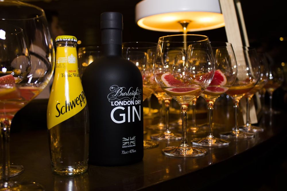 Burleighs London Dry Gin bottle beside Schweppes tonic water with elegant cocktail glasses on a bar.