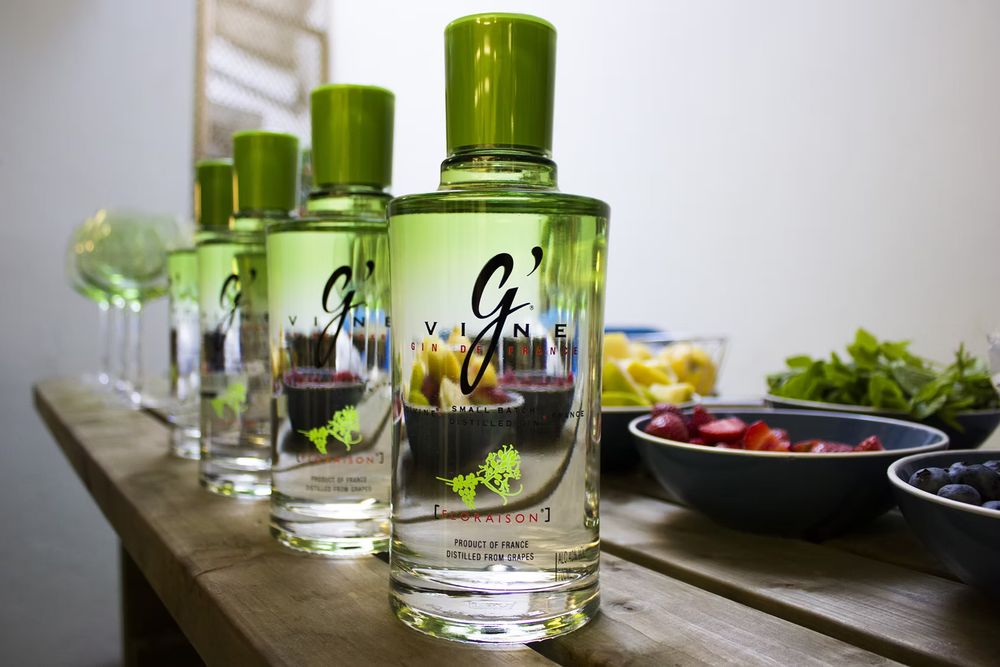 Multiple bottles of G'Vine gin with green caps displayed on a wooden table beside fresh fruits and vegetables.