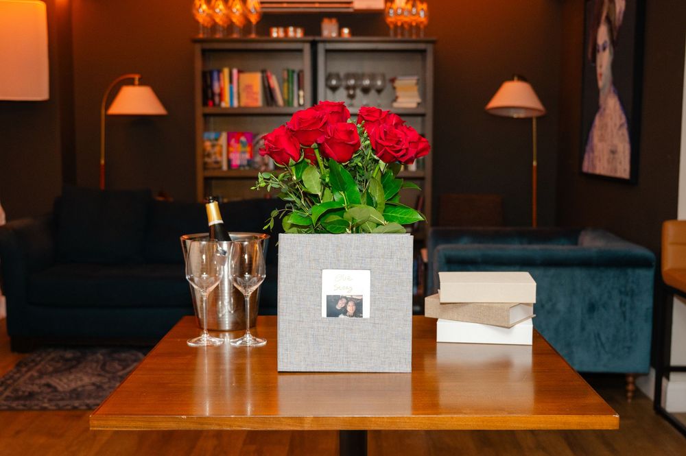 Elegant table with red roses in silver box, champagne glasses, and books in a cozy lounge setting.