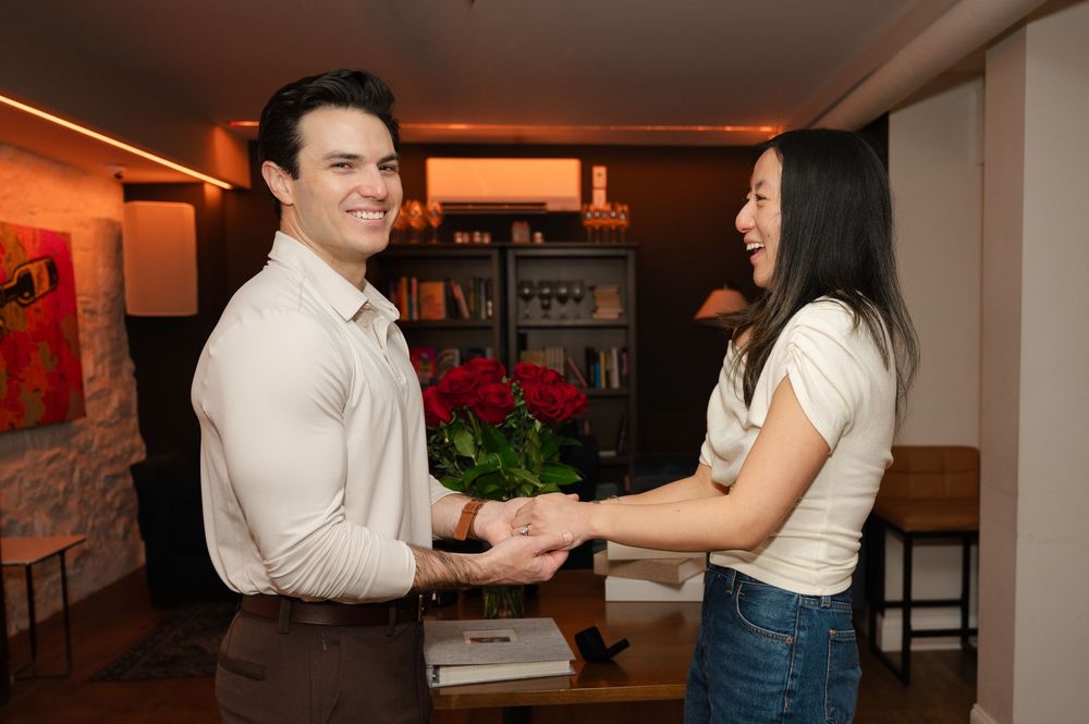 Smiling couple holding hands indoors with a bouquet of red roses in the background.