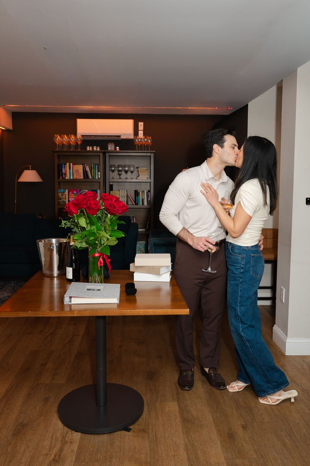A couple shares a romantic kiss beside a vase of red roses and champagne in an elegant room.