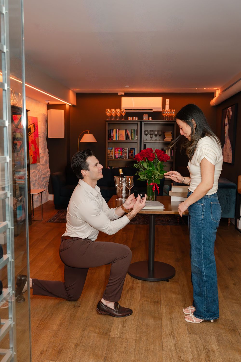 Man proposing on one knee to woman beside a table with red roses and champagne in a warmly lit room.