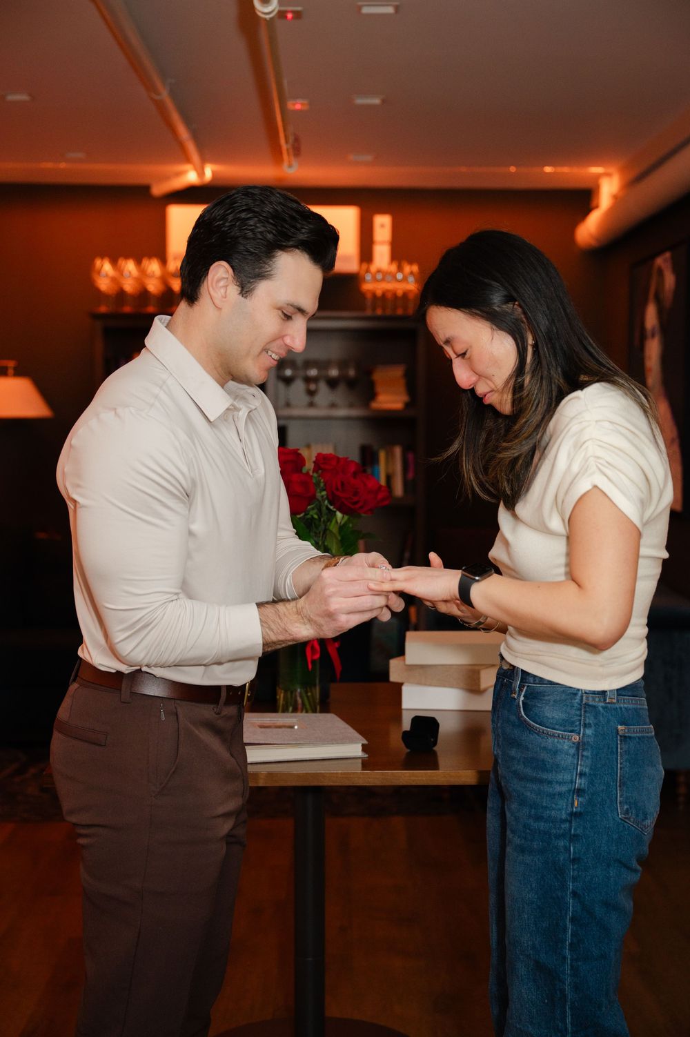 A man places an engagement ring on a woman's finger beside red roses in a warmly lit room.
