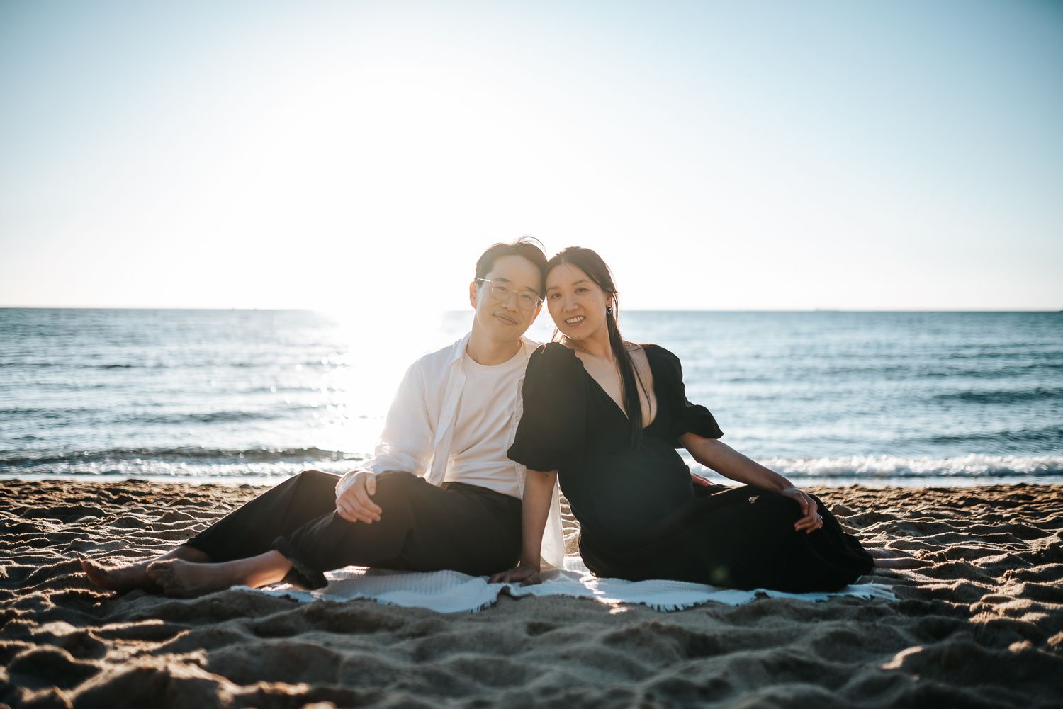 A pregnant couple sitting together on a sandy beach at sunset with ocean waves in the background taken by Melbourne maternity photographer.