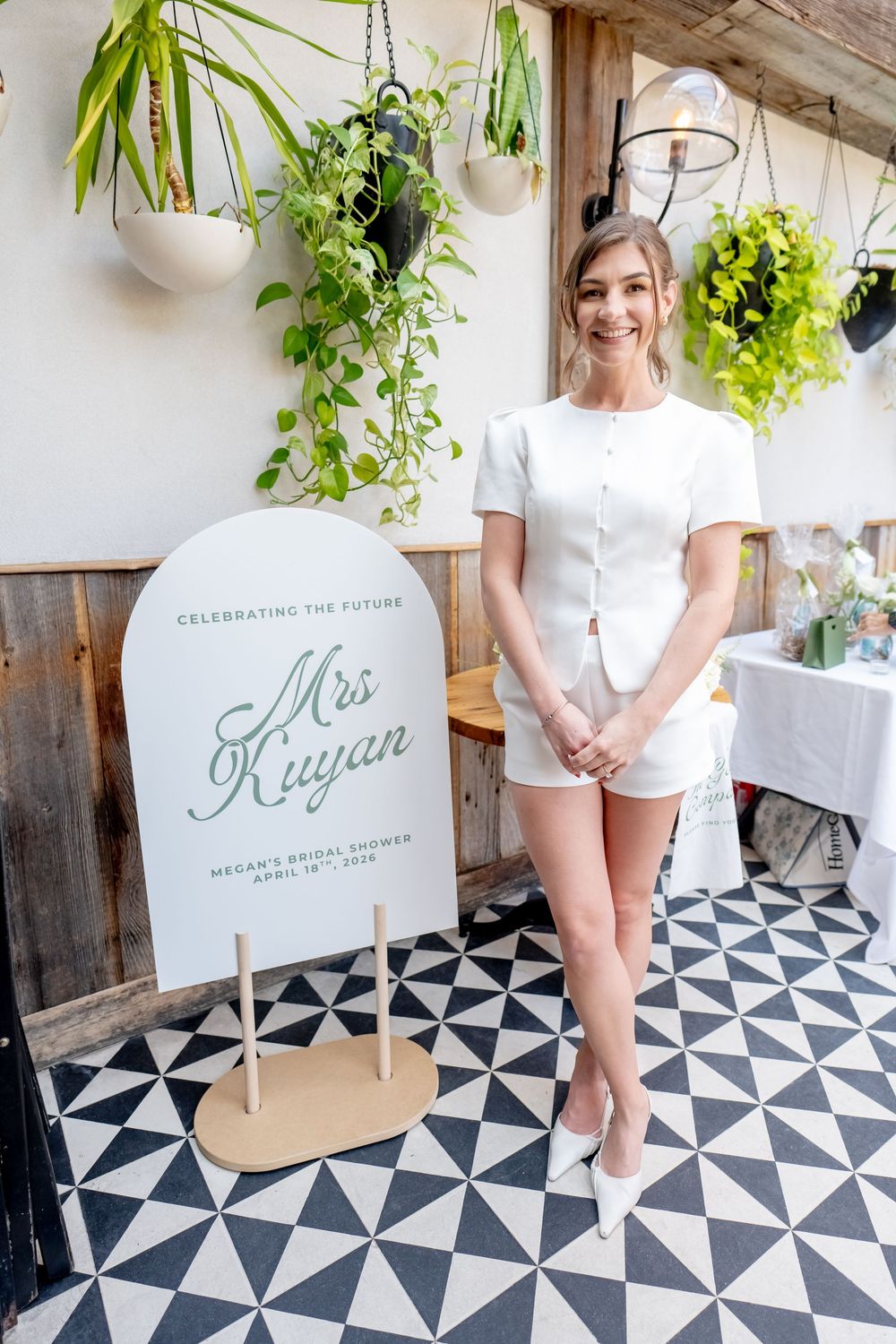 Woman in white romper smiling beside a bridal shower sign reading Mrs. Kuyan, surrounded by lush greenery.