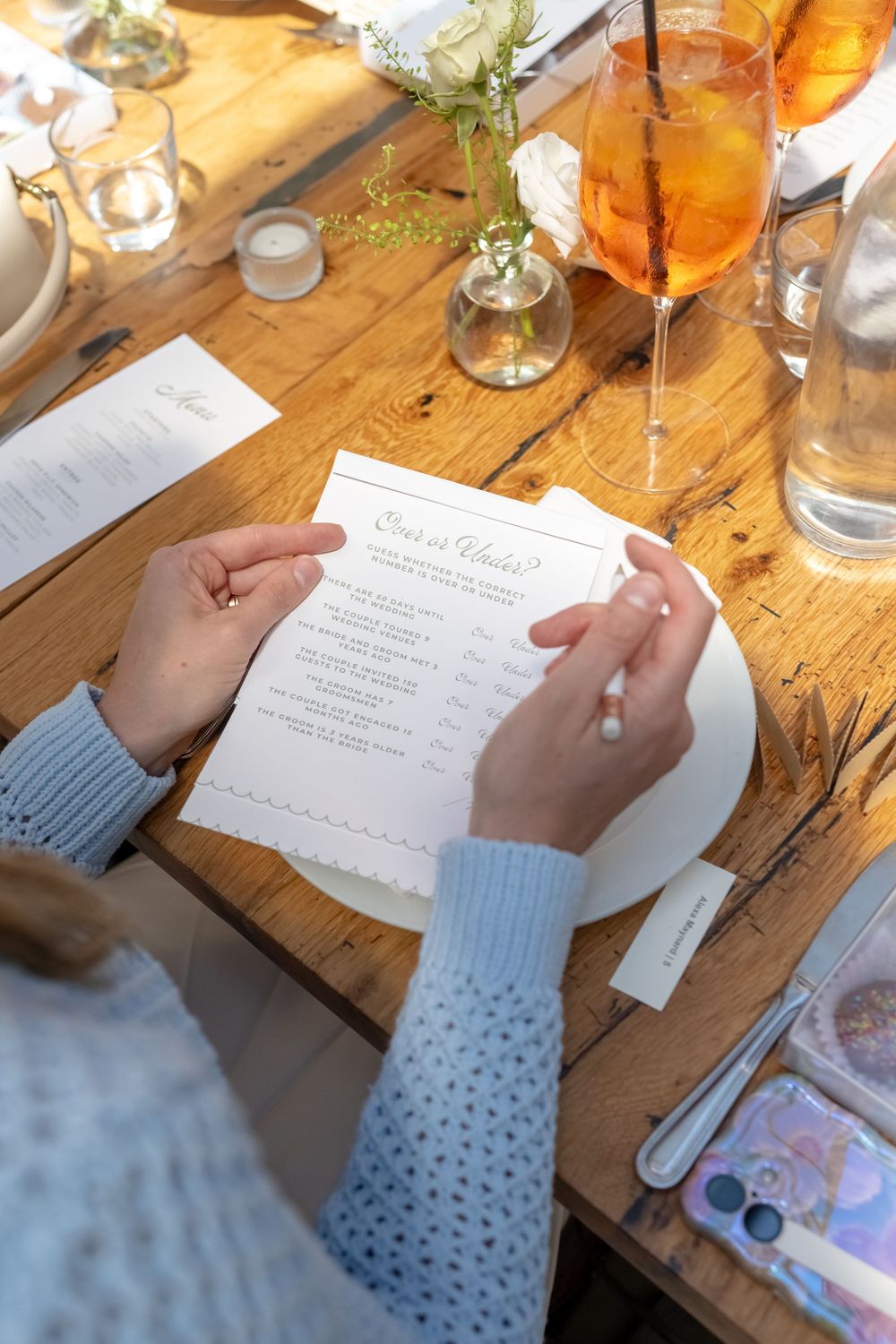 Woman reading a wedding quiz card at a rustic wooden table with candles and an Aperol Spritz cocktail.