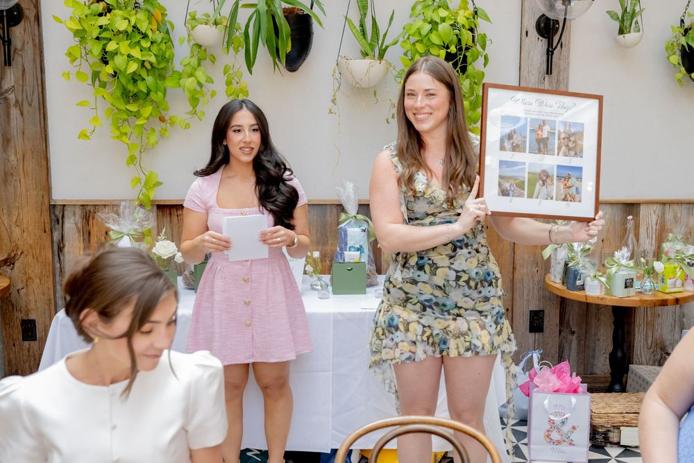 Two women stand at a decorated table holding papers and a framed picture at an indoor garden party event.