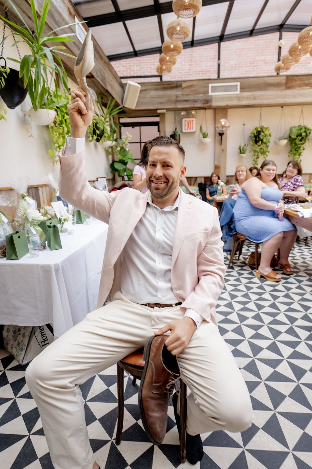 Smiling man in pink blazer raises hand while seated at a chair in a decorated restaurant venue.