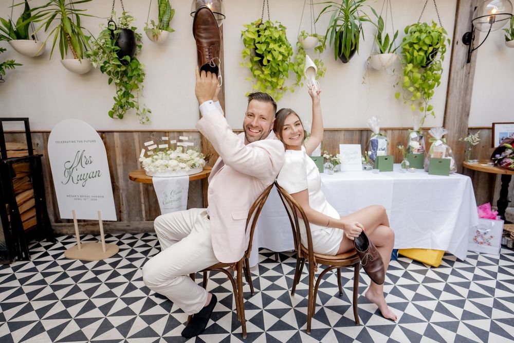 Joyful couple posing back-to-back on chairs at their wedding reception, surrounded by lush hanging plants.