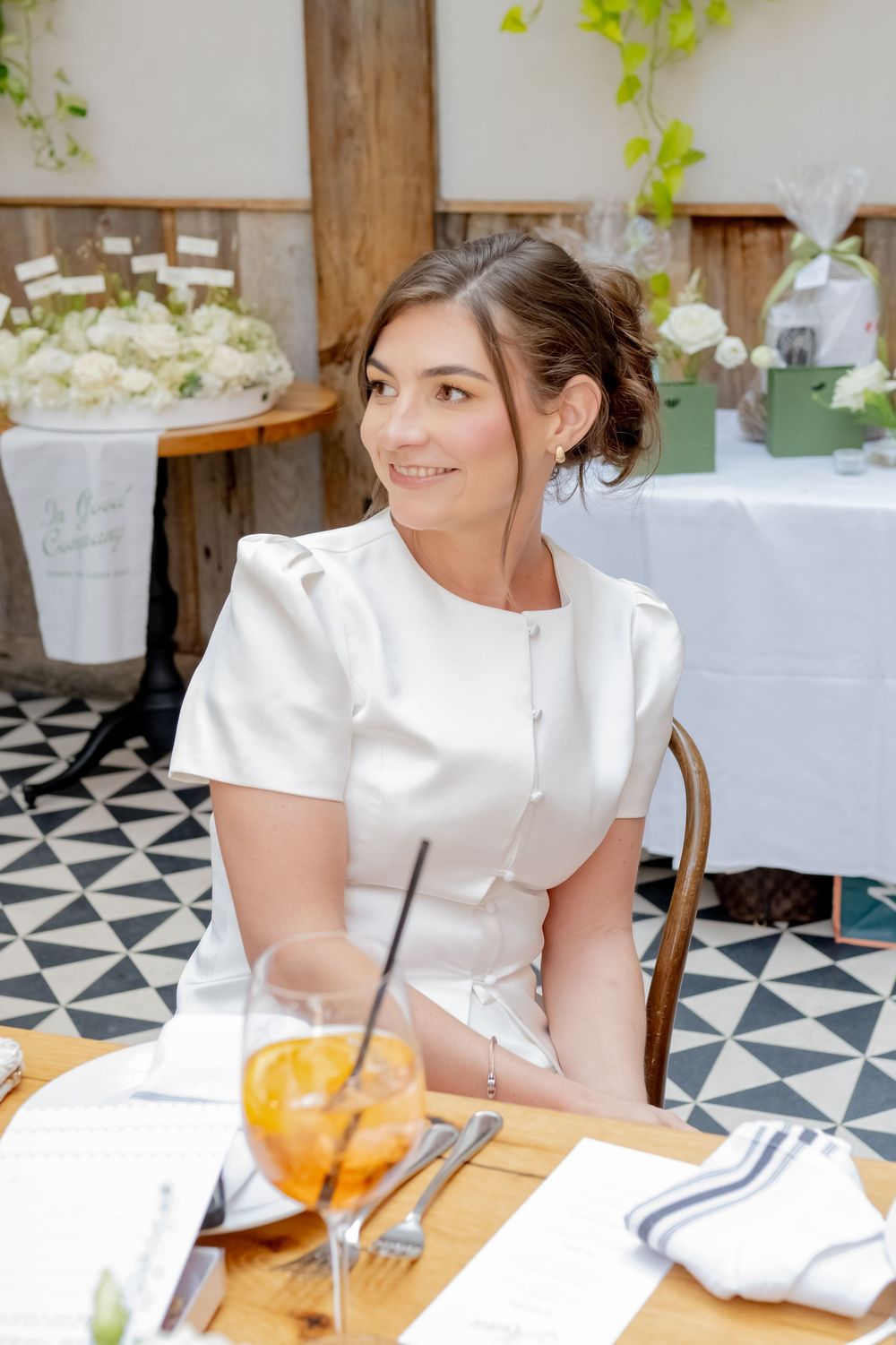 A smiling bride in a white dress sits at a decorated table with floral arrangements and a cocktail at an elegant venue.