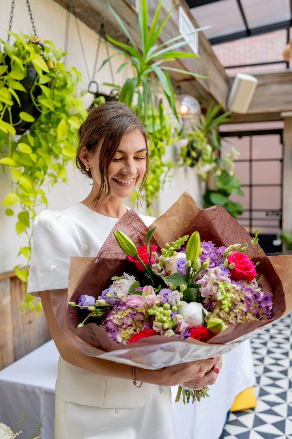 Smiling woman in white dress holding a large colorful bouquet of flowers in a plant-filled indoor setting.
