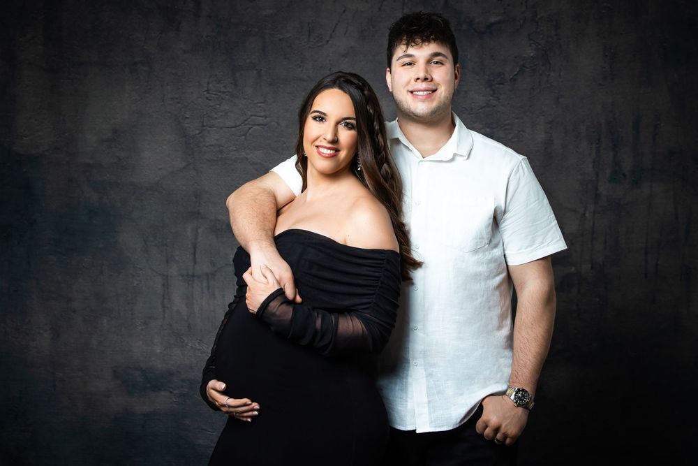 Smiling pregnant woman in black dress with partner during maternity studio photo shoot.