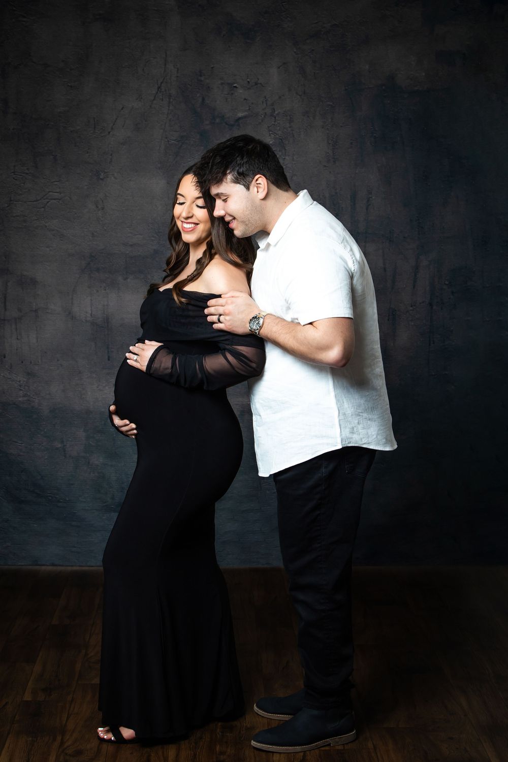 Man tenderly embracing pregnant woman in elegant black maternity gown during studio shoot.