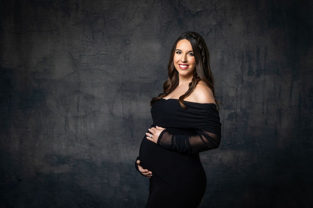 Glowing pregnant woman in off-shoulder black maternity dress smiling during studio photo shoot.