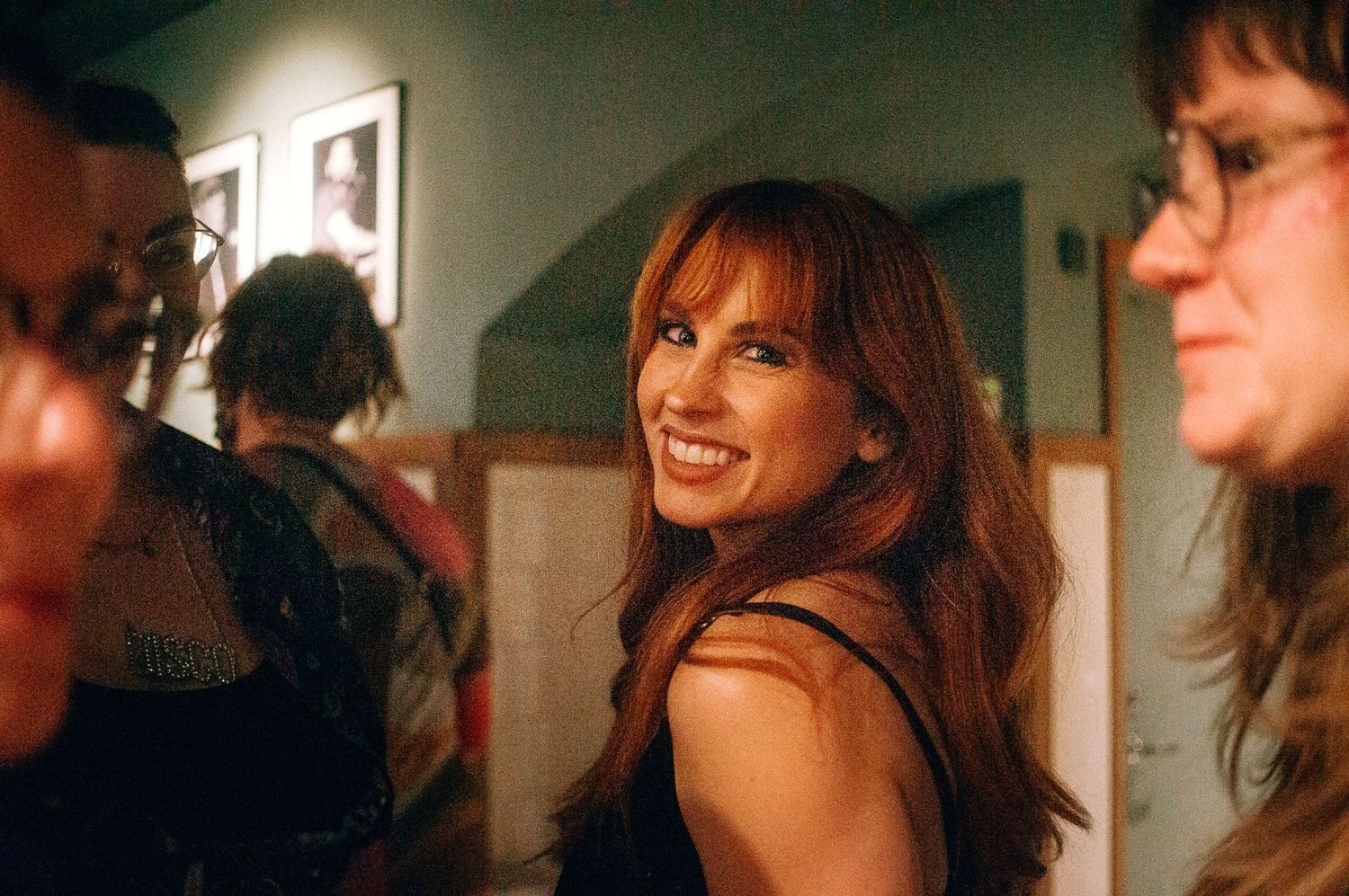 Smiling woman with long auburn hair at an indoor social gathering with people and framed artwork in background.