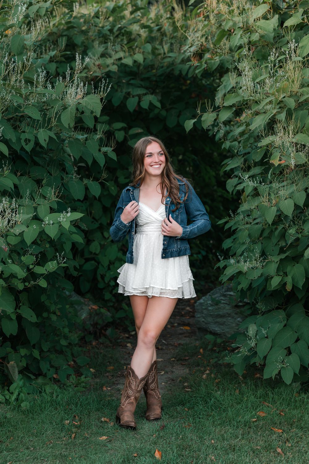 Vermont senior in white dress and denim jacket posing in lush green garden archway with cowboy boots.