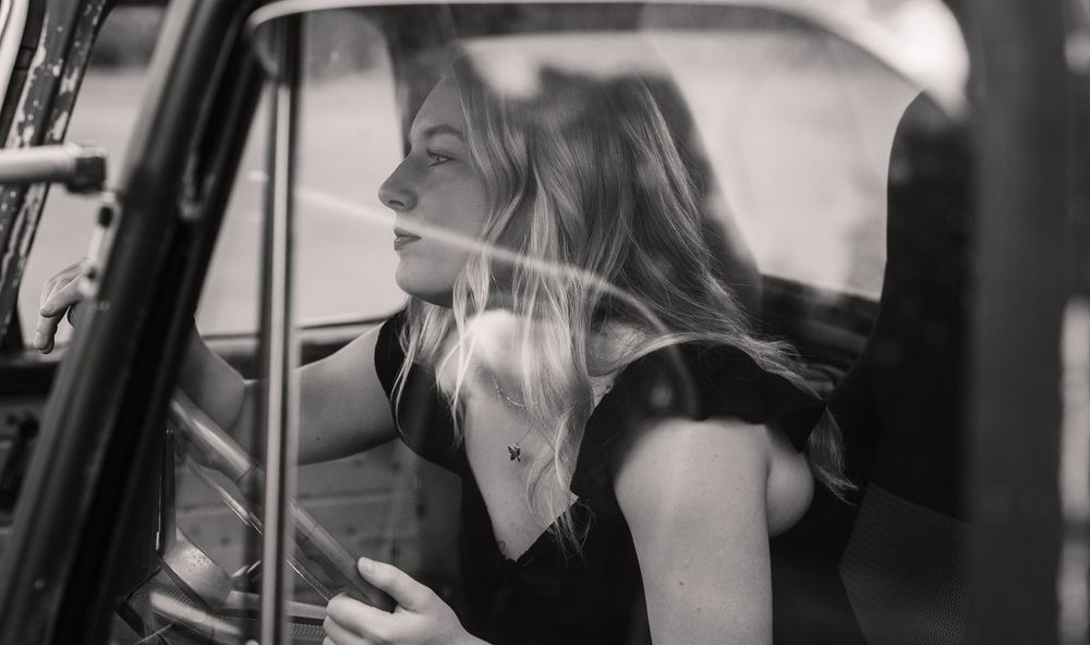 Black and white photo of a blonde woman sitting in a vintage car, gazing out the window with wind-blown hair.