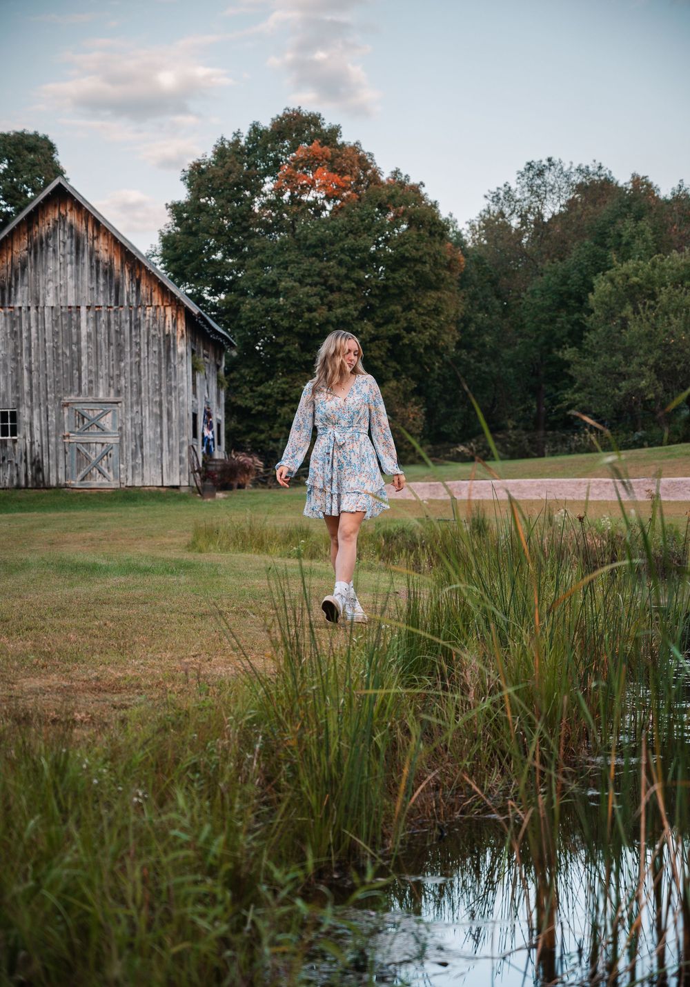A young woman in a floral dress walks near a pond by a rustic barn surrounded by lush green trees.