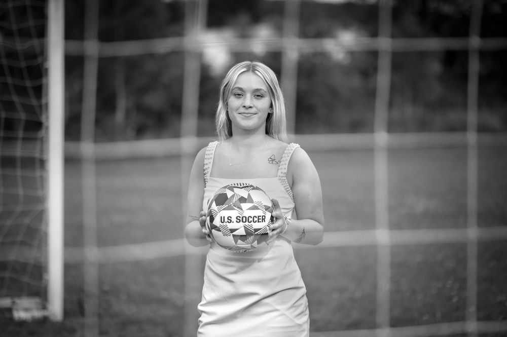 Young blonde woman holding a U.S. Soccer ball in front of a goal net on a soccer field.