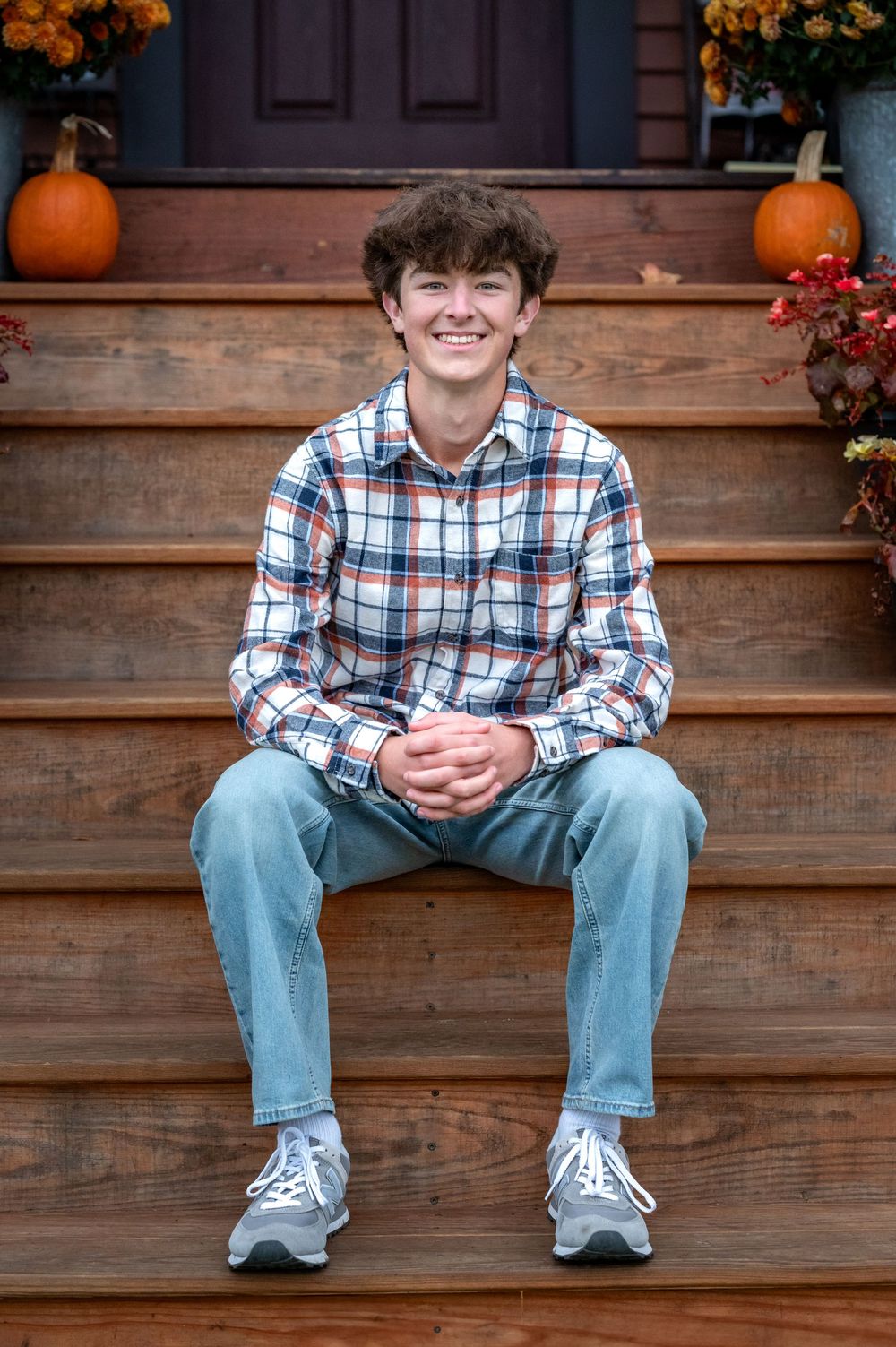 Smiling teen boy in plaid shirt and jeans sitting on wooden porch steps with pumpkins and fall flowers.