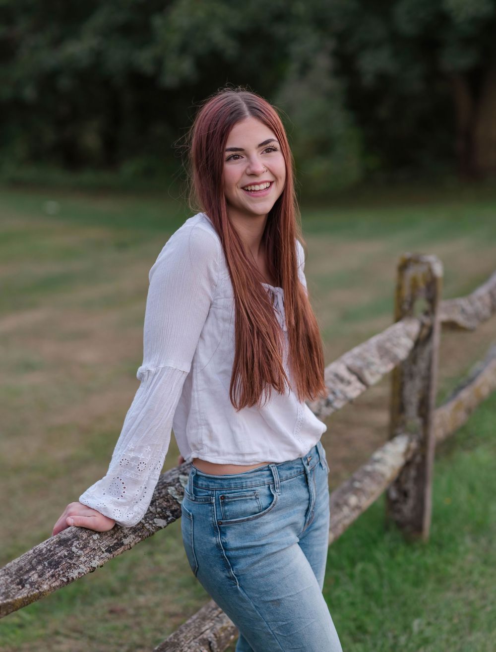 Smiling teenage girl with long red hair leaning on a wooden fence outdoors in jeans and white top.