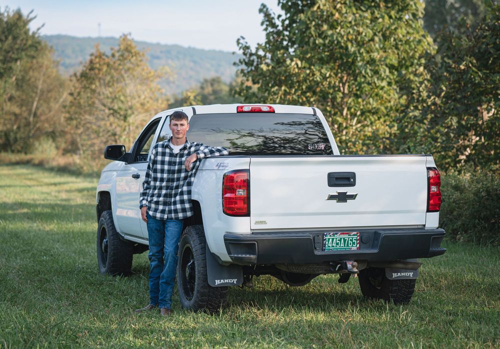 Young man leaning on a white Chevy pickup truck in a green field.