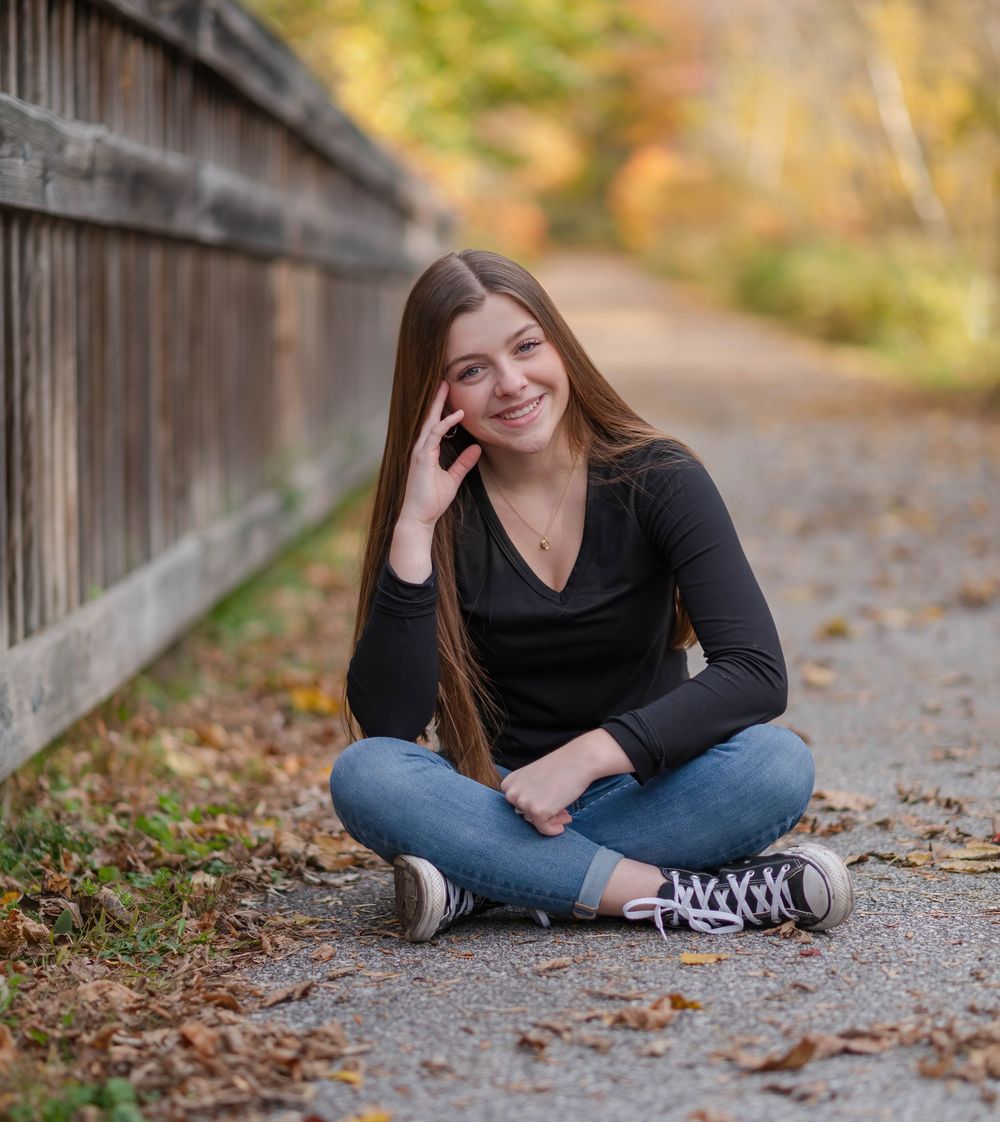 A smiling teenage girl sits cross-legged on a path in jeans and black top, surrounded by autumn foliage.