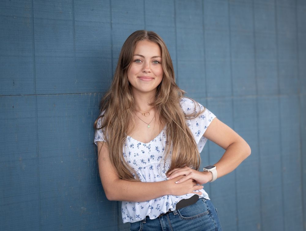 Smiling teenage girl in floral top and jeans posing against a blue wall.